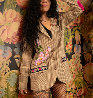Model standing in front of a lush floral wallpapered backdrop wears a beige and green micro-plaid oversized blazer with bodice appliqué and pocket trim, brooches attached to the left lapel, and pink velvet elbow patches, paired with black fishnet stockings