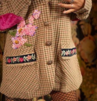 Model standing in front of a lush floral wallpapered backdrop wears a beige and green micro-plaid oversized blazer with pink floral sequined bodice appliqués, embroidered pocket trim, and pink velvet elbow patch, paired with black fishnet stockings