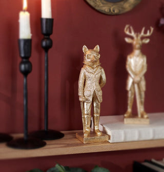 Gold fox and deer statues sit on a bookshelf with taper candles against a red wall