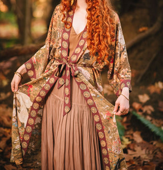 Model standing in an autumnal forest wears a long robe with an art nouveau print and contrasting border and tied belt