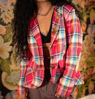 Model wears a pink and multicolored blazer with two mismatched button closures and two gold brooches on the lapel