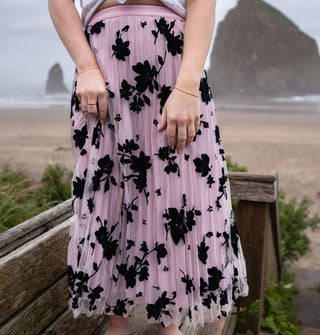 Model wears a pleated pink tulle skirt with black floral embroidery on a beach boardwalk