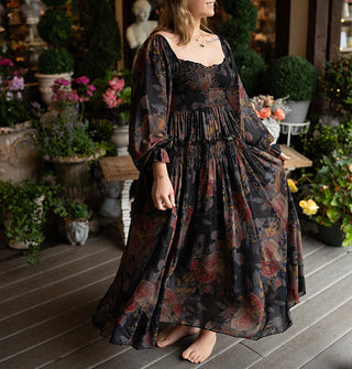 Smiling model twirls in a long-sleeved full-length dark rose print dress with smocked and ruffled details against a decorative porch backdrop