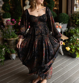 Model twirls in a long-sleeved full-length dark rose print dress with smocked and ruffled details against a decorative porch backdrop