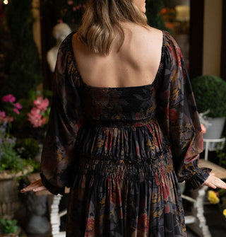 Model facing away from the camera wears a long-sleeved dark rose print dress with smocked and ruffled details and a partially open square back against a decorative porch backdrop