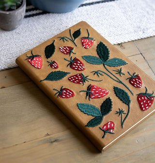Strawberry embroidered brown hardcover journal rests on a wooden tabletop set with linen and potted plants