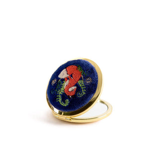 Opened round compact mirror with gold rim features a dark blue cover with embroidered design of a fox flanked by curling fern tendrils