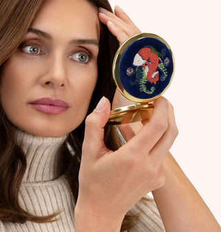 Model holds a round compact mirror with gold rim that features a dark blue cover with an embroidered design of a fox flanked by curling fern tendrils