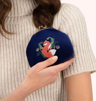 Model wearing a ribbed beige turtleneck sweater holds a dark blue velvet kiss lock change purse with an embroidered design of an orange and white fox between delicate curling fern tendrils