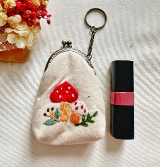White linen pouch next to a tube of lipstick features a gunmetal kiss lock closure with attached keychain and colorful embroidered mushroom design on the fabric body