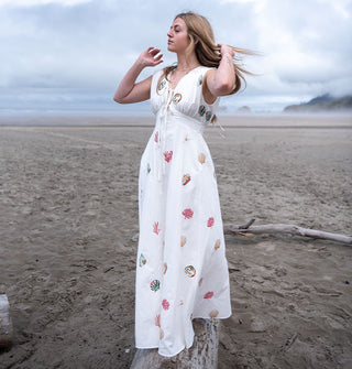 Model standing on driftwood with hair blowing in the wind wears a white maxi-length A-line dress embroidered with colorful seashells and coral on a misty beach