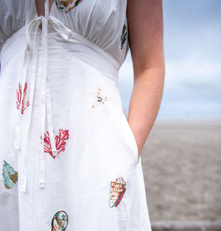 Model wears a white A-line dress embroidered with colorful seashells and coral on a misty beach
