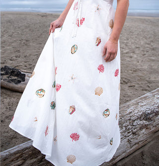Model standing on driftwood wears a white maxi-length dress embroidered with colorful seashells and coral on a misty beach