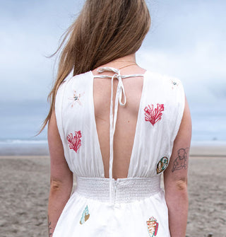 Model wears a white dress with tied open back and colorful seashell and coral embroidery on a misty beach