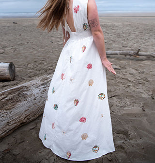 Model standing beside a piece of driftwood with hair blowing in the wind wears a white maxi-length open-back  dress embroidered with colorful seashells and coral on a misty beach