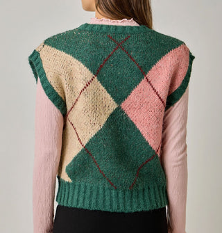 Model facing away from the camera wears a green, beige, pink, and burgundy argyle sweater vest with cap sleeves and ribbed hems over a pink lettuce-edged mock turtleneck