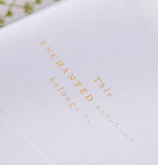 Closeup of a notebook's inside page features the words, "This enchanted notebook belongs to:" in gold lettering