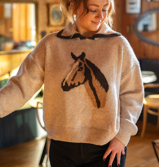 Smiling model wears a beige sweater with a central brown and blue horse head design and a wide collar with dark blue edge