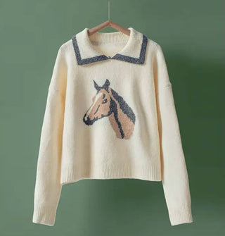 Cream-colored sweater with gray-trimmed collar and central horse head design in gray, brown, and white hangs in front of a green backdrop
