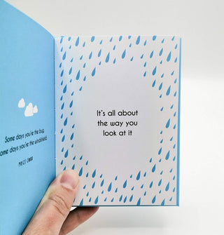 Model's hand holds the Every Cloud Has a Silver Lining book open to a page that says, "It's all about the way you look at it" surrounded by blue raindrops