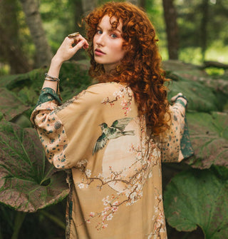 Model turned slightly away from the camera with arm raised wears a printed robe with bird and blossoms designs in a lush botanical setting