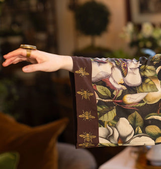 Model outstretched arm wears the wide sleeve of a robe with large, lush floral pattern and brown trim with gold bees