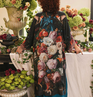 Model standing at a lushly decorated table facing away from the camera wears a robe with a dramatic floral and hummingbirds design on the back