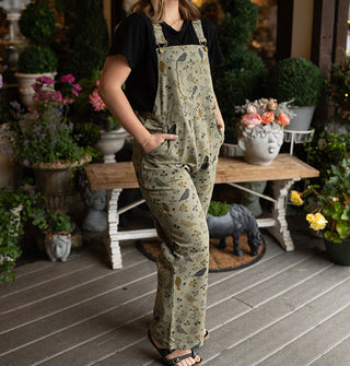 Model poses with hands in the pockets of olive green bib overalls with birds and branches print on a decorative porch deck