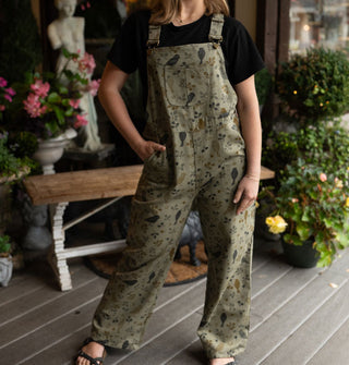 Model poses with hand in the pocket of olive green bib overalls with birds and branches print on a decorative porch deck