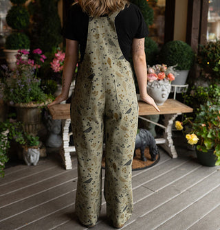 Model facing away from the camera wears olive green bib overalls with birds and branches print on a decorative poch deck