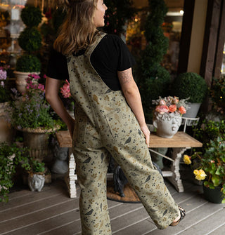 Smiling model wearing olive green bib overalls with birds and branches print kicks out one leg