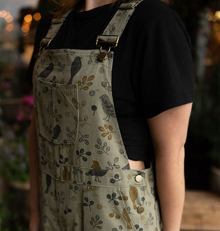 Model wears olive green bib overalls with birds and branches print