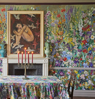 Dense, colorful floral print wallpaper is applied to a dining room with fireplace, fine art, and a dining table with matching tablecloth topped with a candelabra