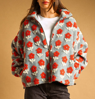 Model poses with hands in the pockets of a light blue sherpa jacket featuring all-over flocked orange and white flowers, a full-length zipper, collar, and side toggles