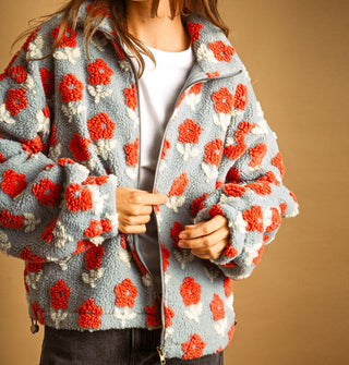 Model wears a light blue sherpa jacket featuring all-over flocked orange and white flowers, a full-length zipper, collar, and side toggles