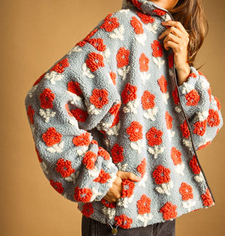 Model poses with hand in the pocket of a light blue sherpa jacket featuring all-over flocked orange and white flowers, a full-length zipper, collar, and bottom side toggle
