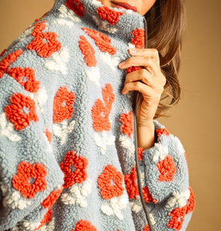 Model pulls at the collar of a light blue sherpa jacket with bold, all-over orange and white flower pattern