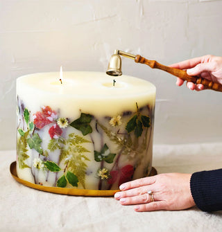 Model snuffs one of two wicks on a large botanical candle featuring various leaves, ferns, and flowers embedded in its wax and positioned on a gold platter against a roughly plastered white backdrop
