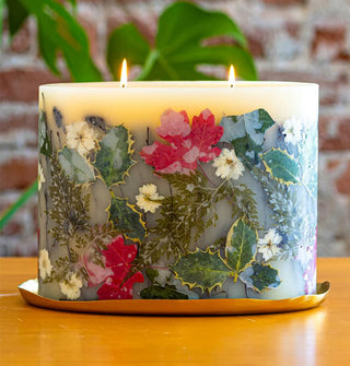 Lit two-wick botanical candle with various leaves, flowers, and ferns embedded in its wax