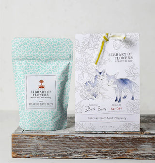 Inner and outer pouches of Library of Flowers Forget Me Not Bath Salts featuring monochromatic floral and fox arwork rest on a rustic wooden platform