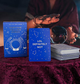 A fortune teller poses with a crystal ball and blue Fortune Telling Cards box and sample card reading, "LOL definitely not" staged on a purple velvet-covered tabletop