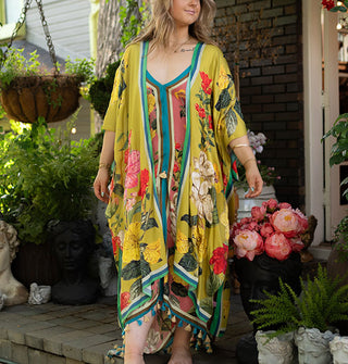 Model wears a long olive-chartreuse kimono-style robe with large floral print and contrast edge stripes paired with a dress with tassels along the bottom