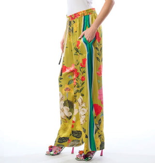Model poses with hand in the pocket of a pair of wide-legged chartreuse pants featuring a large floral print, side seam contrast stripes, and an elastic waistband with ties