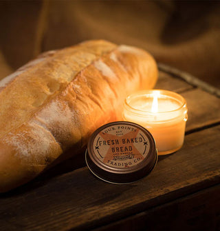 Lit Fresh Baked Bread jar candle with lid removed is staged on a wooden surface next to a loaf of bread