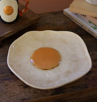 Plate shaped and painted to resemble a fried egg with a yellow yolk center and irregular edge rests on a wooden surface with a matching mug and stack of books
