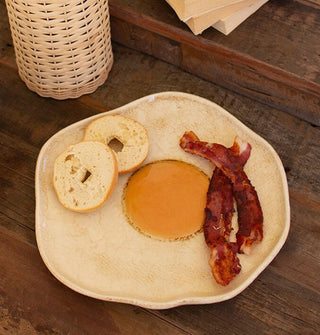 Plate shaped and painted to resemble a fried egg with a yellow yolk center and irregular edge holds a mini bagel and two strips of cooked bacon