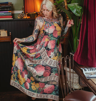 Model poses in a furnished study space wearing a colorful floral maxi-length slip dress that says, "Friendship, Love and Truth" on it in a banner design