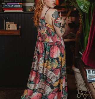 Model poses in a furnished study space wearing a colorful floral maxi-length slip dress that says, "Friendship, Love and Truth" on it in a banner design