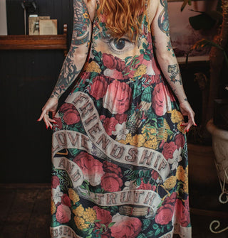 Model poses in a furnished study space wearing a colorful floral maxi-length slip dress that says, "Friendship, Love and Truth" on it in a banner design