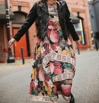 Model poses on a city street wearing a colorful floral maxi-length slip dress that says, "Friendship, Love and Truth" on it in a banner design with black leather jacket overtop